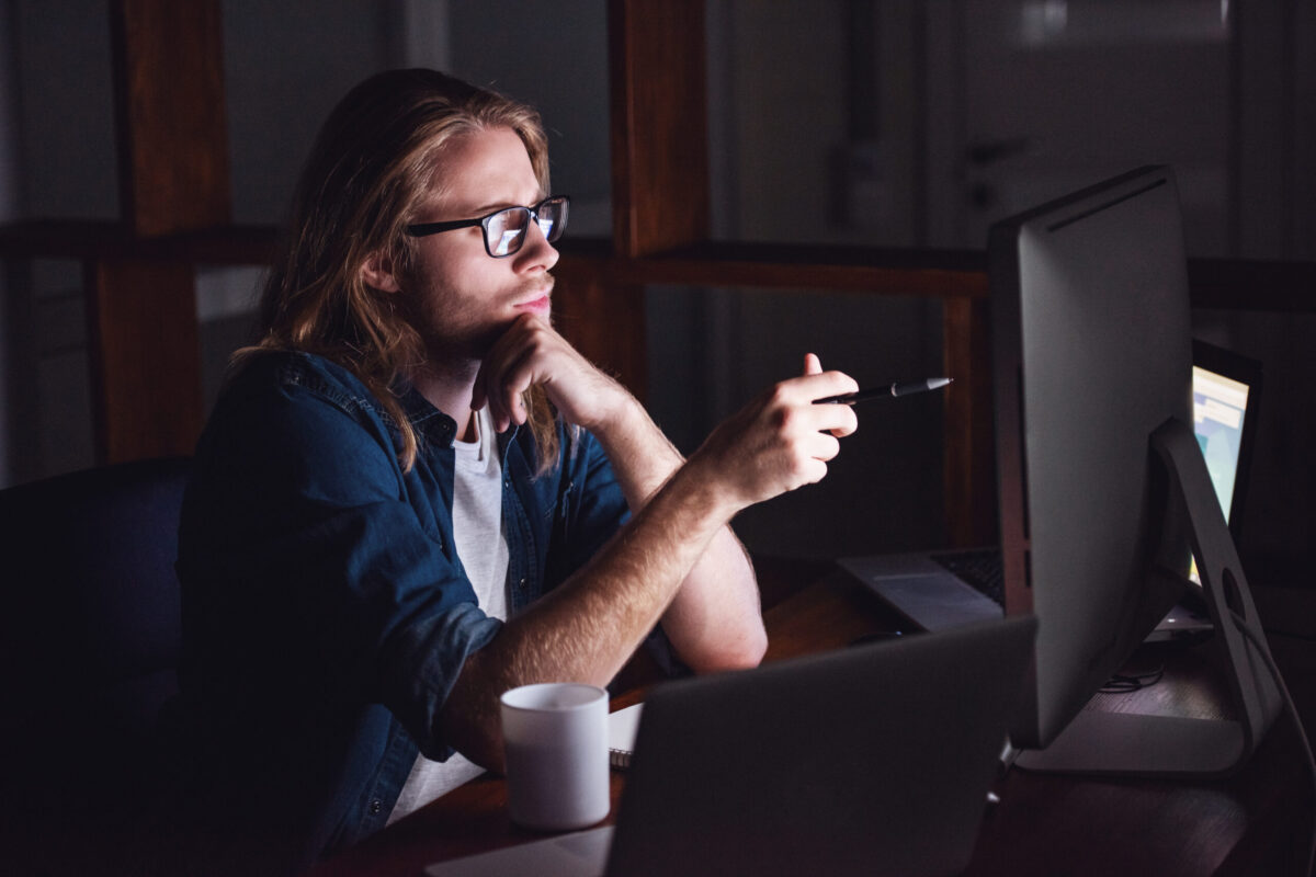 Handsome young businessman with shoulder-length blond hair and in casual wear and eyeglasses is working with a computer at night