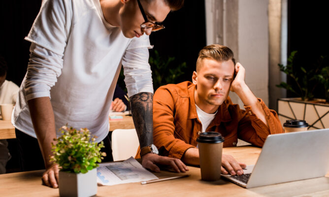 young businessman standing near exhausted colleague using laptop at night in office