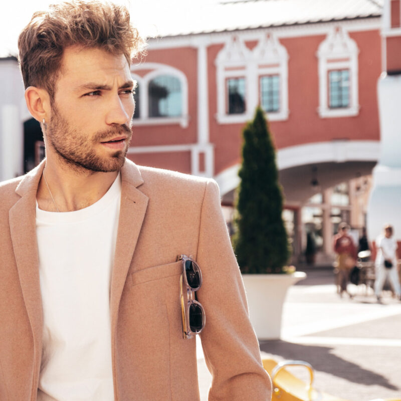 Handsome confident stylish hipster lambersexual model.Sexy modern man dressed in elegant beige suit. Fashion male posing in the street background in Europe city at sunset