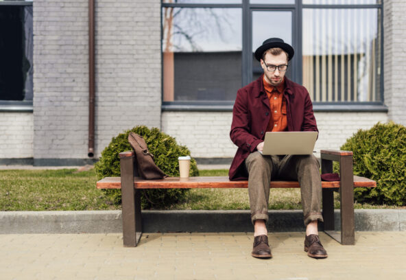 stylish man using laptop and sitting on bench with coffee and bag