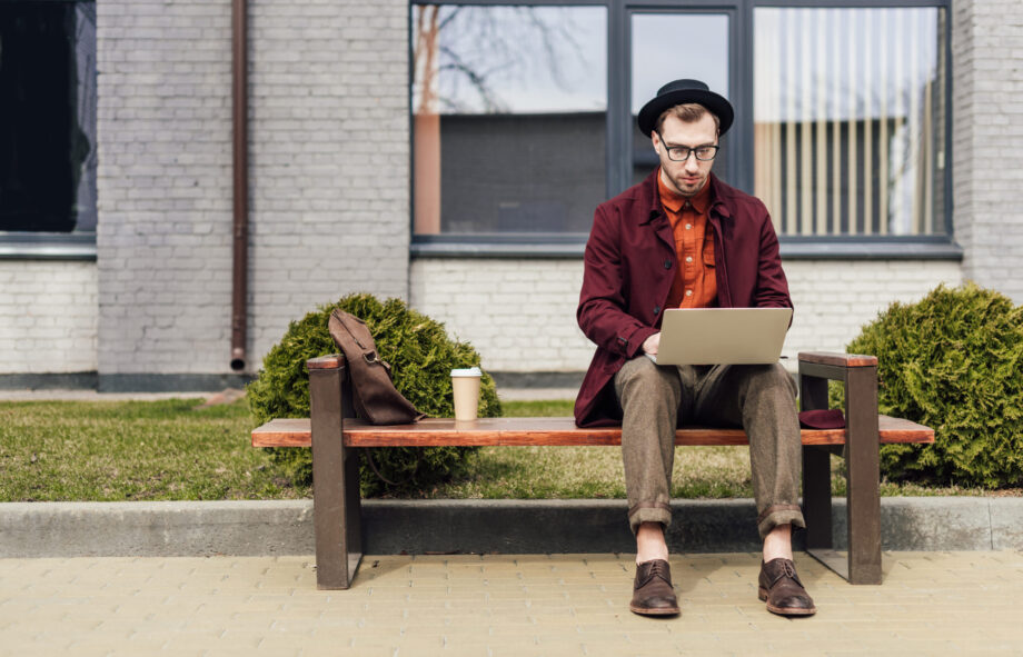 stylish man using laptop and sitting on bench with coffee and bag